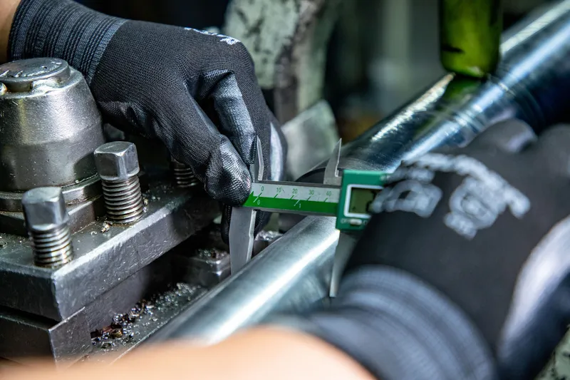 Gloved technician checking a machined steel shaft with a digital caliper